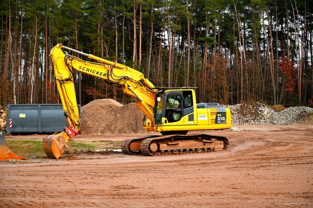 A yellow excavator at work in a forested construction site, surrounded by dirt and gravel.