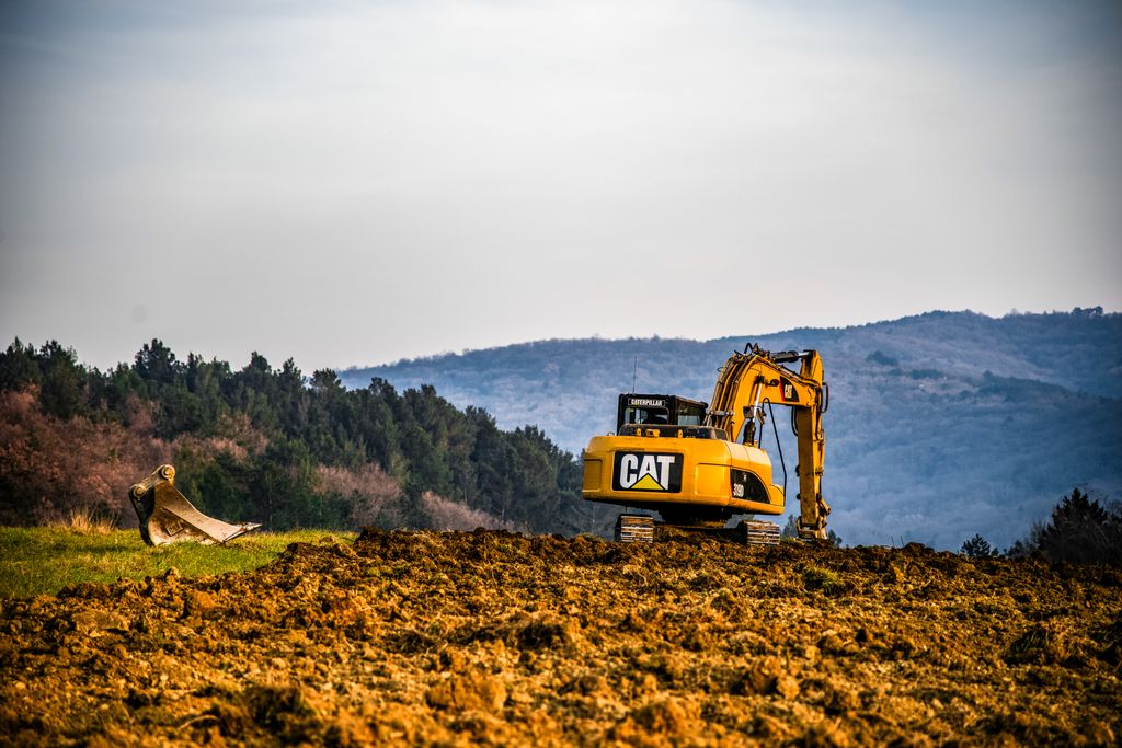 A yellow excavator working on a field with a scenic forest and hills in Dekani, Slovenia.